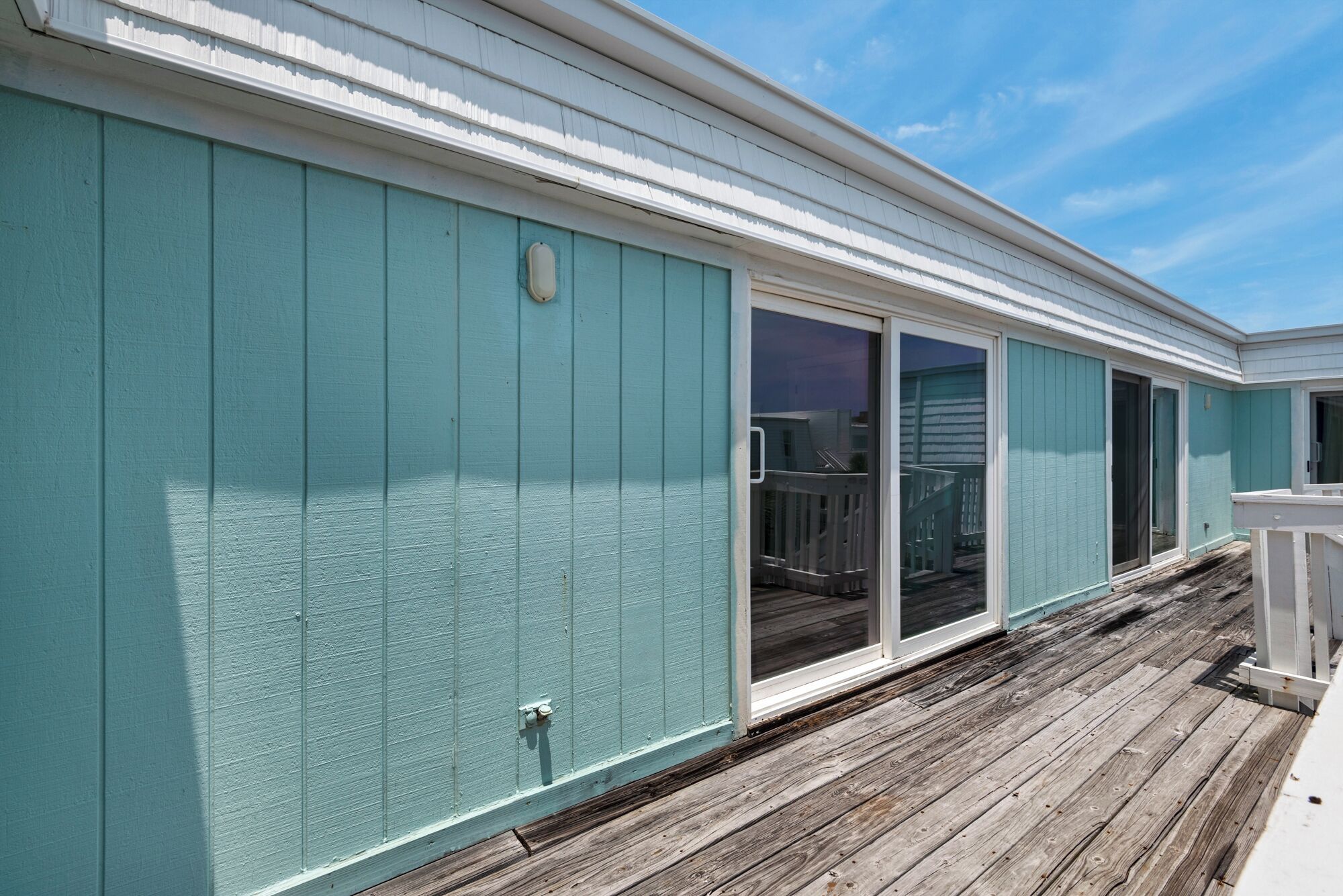 Un bâtiment à la façade de couleur turquoise, doté d’une terrasse en bois et d’une porte-fenêtre coulissante.