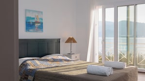 Een slaapkamer met een bed, een nachtkastje, een lamp, een schilderij en uitzicht op zee door een groot raam.