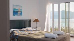 A bedroom with a bed, a nightstand, a lamp, a painting, and a view of the sea through a large window. - Alekos Beach Houses-Aspasia (Kefalonia)