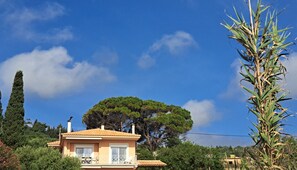 A two-story house with a balcony, surrounded by trees and a clear blue sky. - Alekos Beach Houses-Aspasia (Kefalonia)