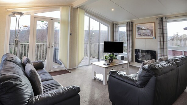 A spacious living room with a large black leather sofa, a flat-screen TV, and a fireplace. - Islay Lodge (Spean Bridge)