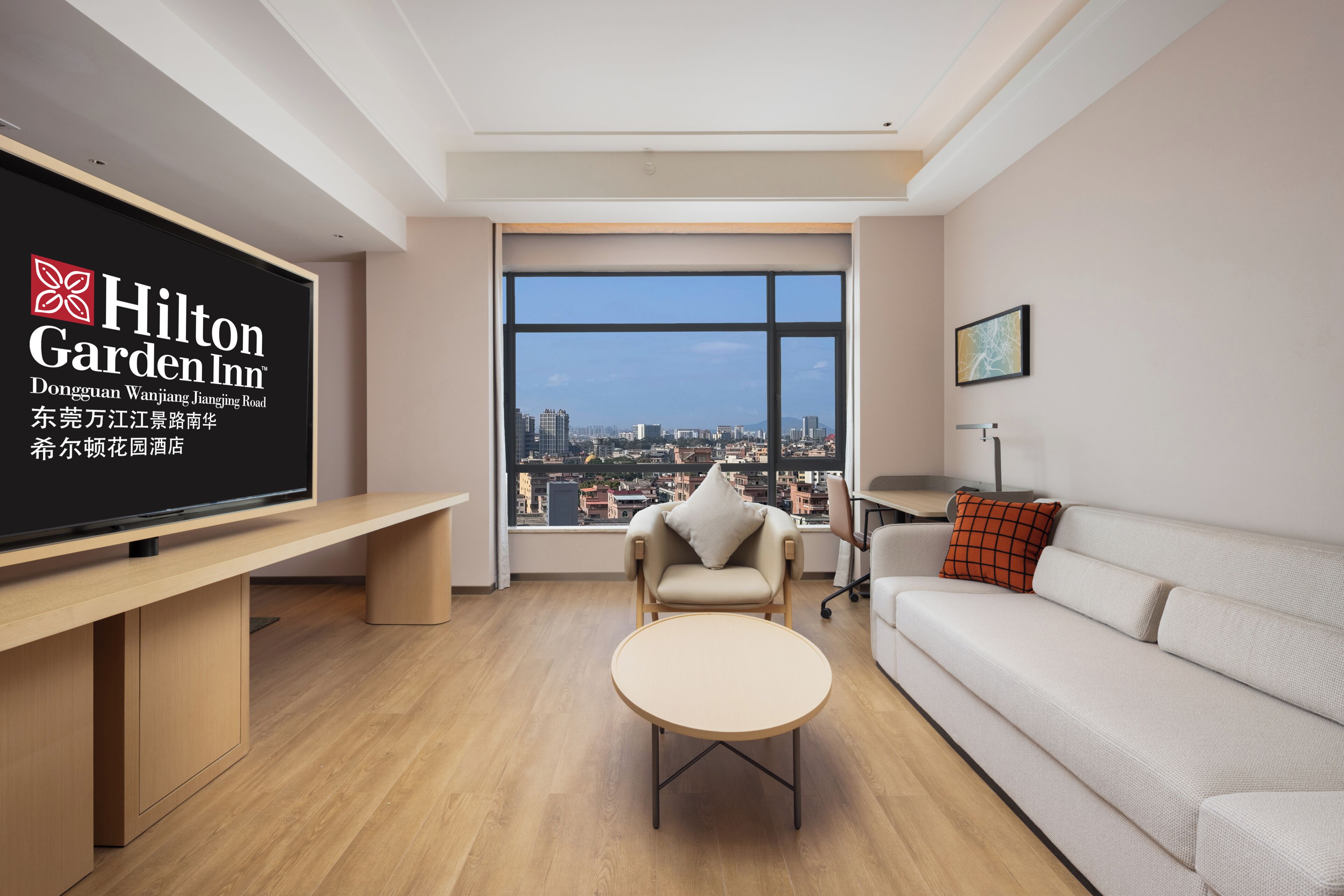 A modern hotel room with a large window, a flat-screen TV, a sofa, a small table, and a chair.