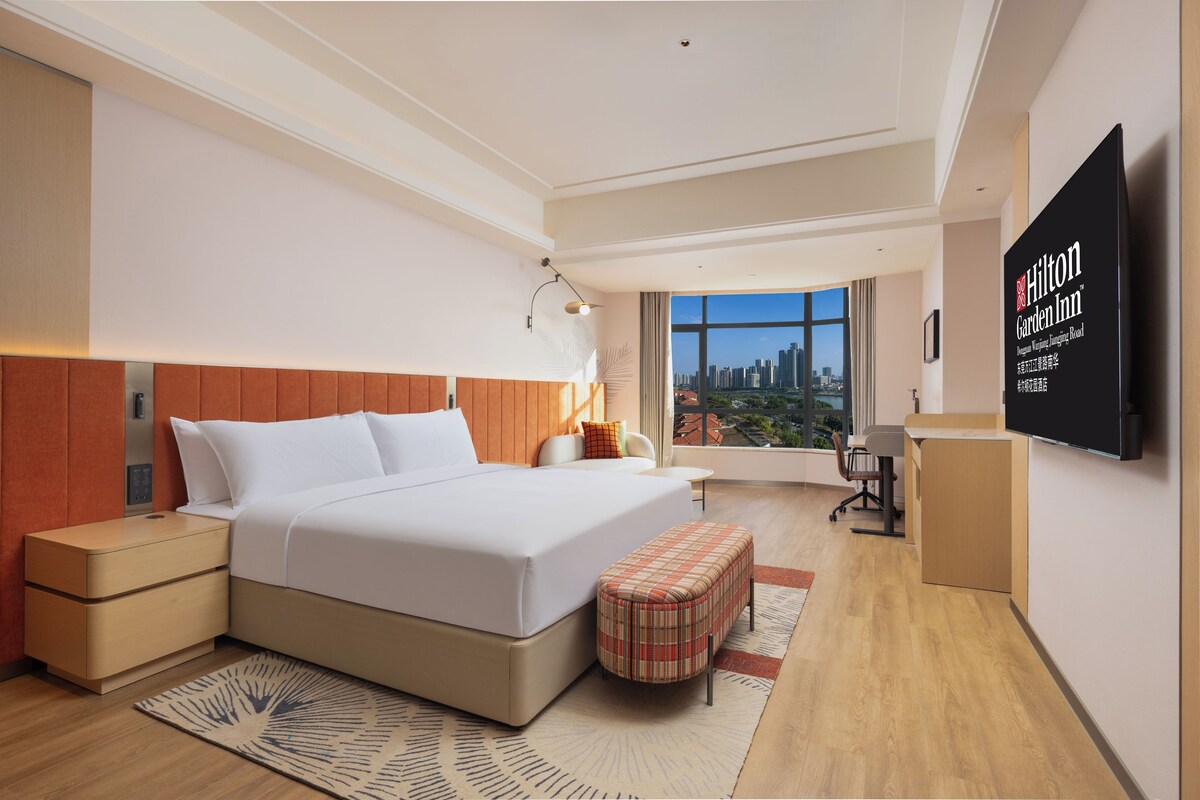 A modern hotel room with a large bed, a desk, a chair, a television, and a city view.