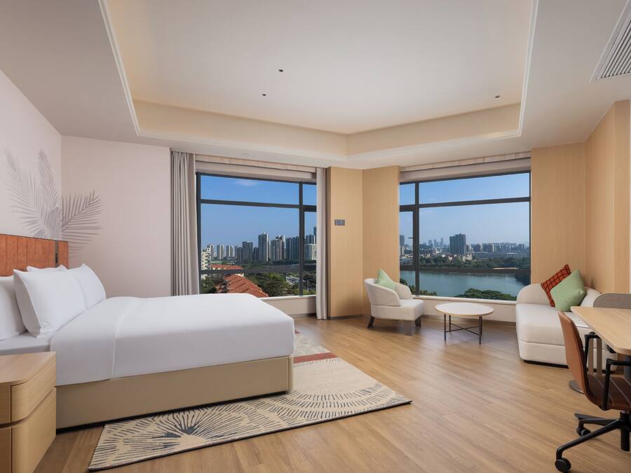 A modern hotel room with a large bed, a seating area, a dining table, and large windows offering a city view.
