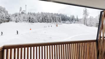 Blick auf eine verschneite Skipiste durch ein Fenster mit Holzlamellen.