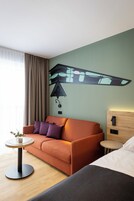 Una habitación de hotel con sofá, una mesita redonda con un jarrón, cama, luz adosada a la pared y cabecero de madera.