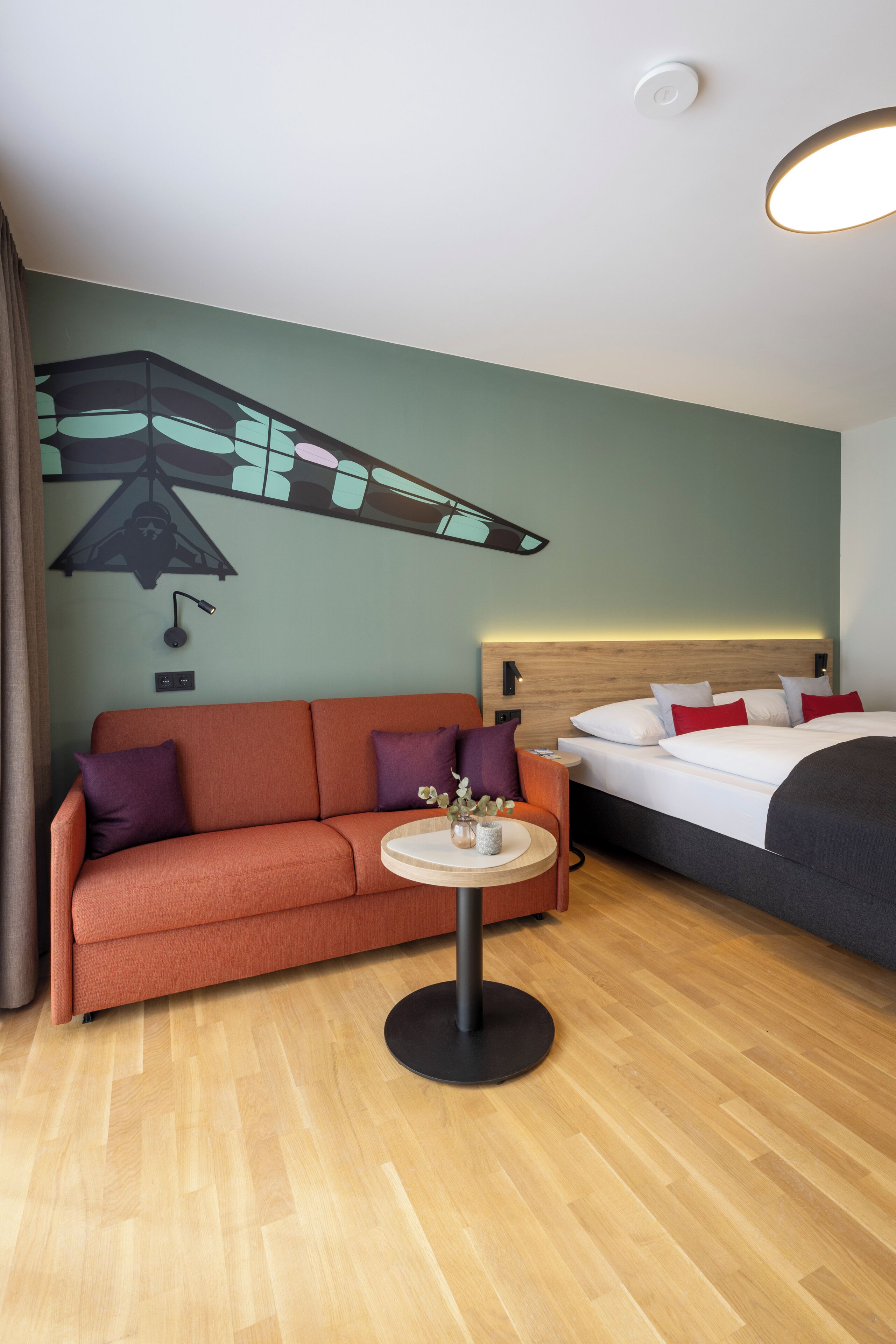 A hotel room with a sofa, a small round table, a bed, and a wall mural.