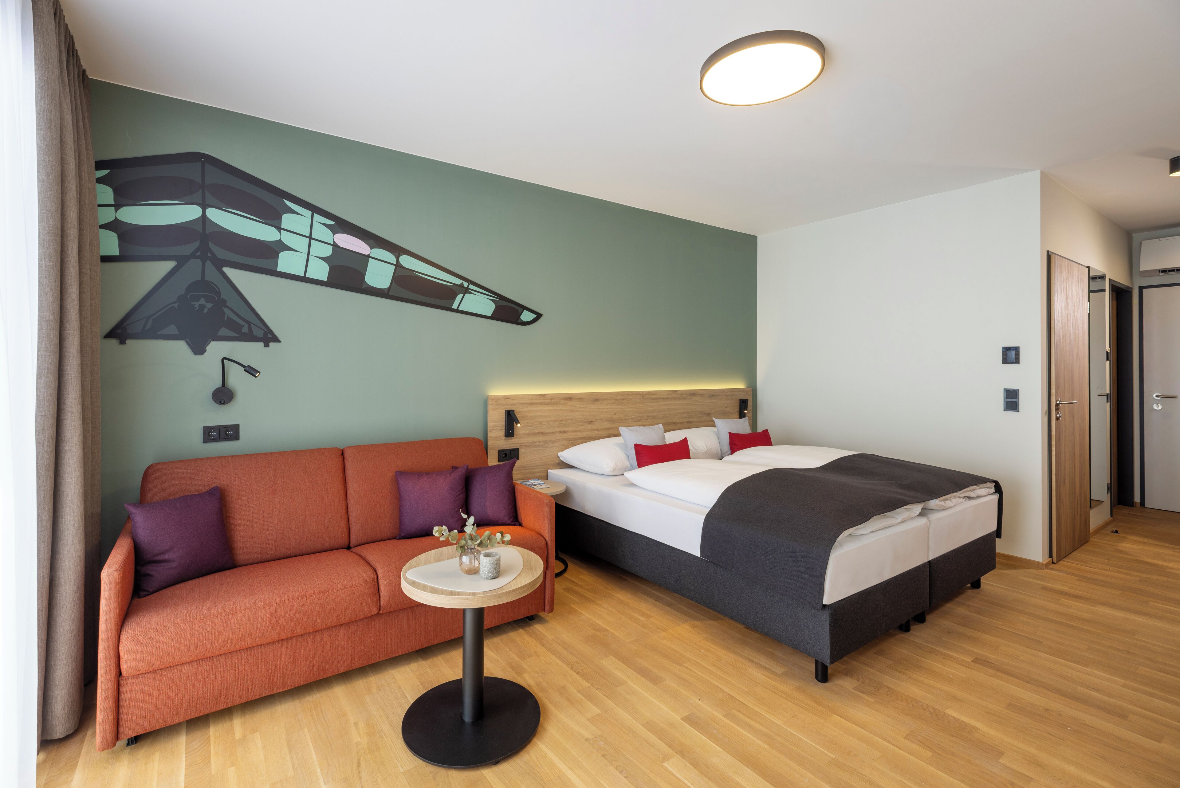 A hotel room with a bed, a sofa, a small round table with a vase, and a wall mural.