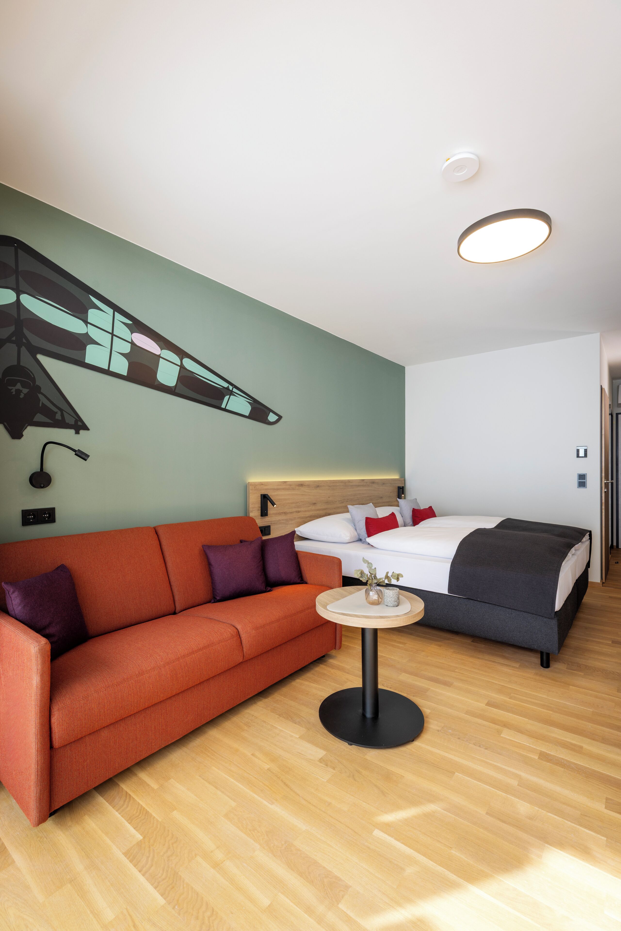 Una habitación de hotel moderna con suelo de madera, un sofá, una cama con cabecera, una mesita redonda pequeña y una pared con diseño geométrico.