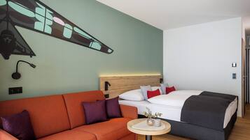 Une chambre d’hôtel moderne avec un plancher en bois, un canapé, un lit avec une tête de lit, une petite table ronde et un mur orné d’un motif géométrique.