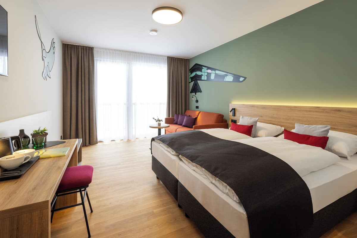 Een hotelkamer met een bed, een bureau met stoel, een bijzettafeltje met plant en een raam met gordijnen.