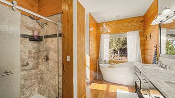 A bathroom with a shower, a bathtub, a vanity with a mirror, and a window with a view of trees.