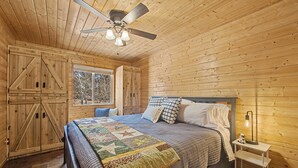 Un dormitorio de cabaña de madera con una cama, colcha, ventilador de techo y una mesita de noche con lámpara.