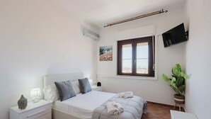A bedroom with a bed, bedside table, lamp, air conditioning unit, window, and a potted plant.