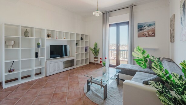 A modern living room with a flat-screen TV, white shelving units, a grey sofa, and a large window with a view of the city.