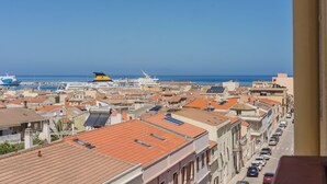 A coastal cityscape with a view of the sea, featuring traditional buildings with red-tiled roofs, a marina with boats, and a clear blue sky.
