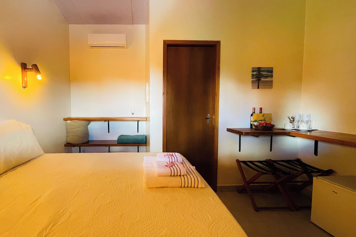 Ein Hotelzimmer mit einem Bett, einer Bank, einem kleinen Tisch mit Flasche und Gläsern sowie einer Tür.