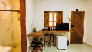 A compact room with a wooden desk, a television, a small refrigerator, and a window with wooden shutters. - Vila Bonita Guest House (Alto Paraíso de Goiás)