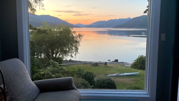 A room with a large window overlooking a lake and mountains during sunset. - Waterfront Vacation Home -only for longer term stays(please contact for details) (Sechelt)
