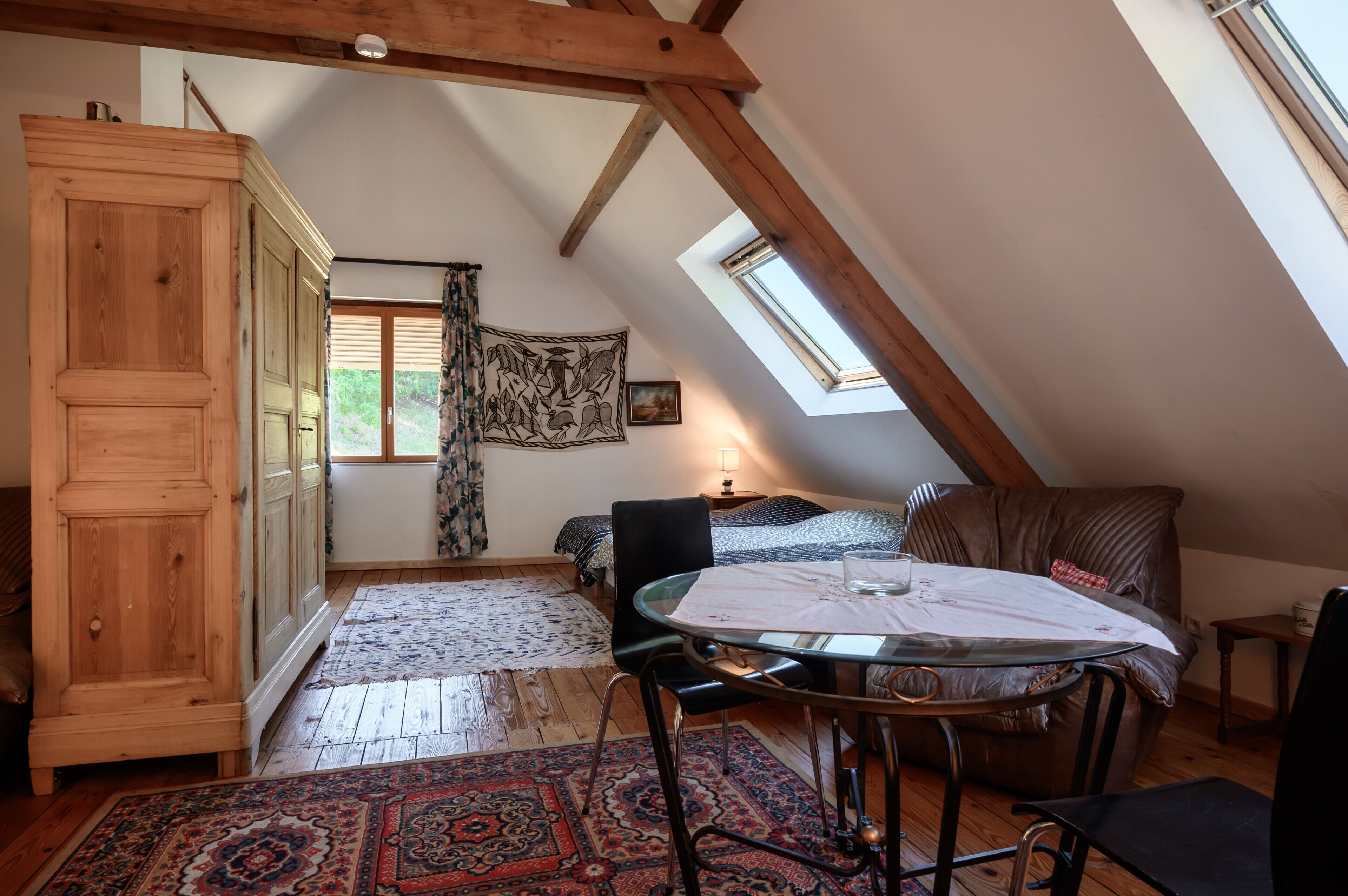 Et hyggeligt soveværelse på loftet med skråvægge, en stor garderobe, en seng med et mønstret sengegavl, et lille bord med et glas, og udsigt fra et vindue.