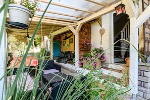 Un área de estar al aire libre techada, con mobiliario de mimbre, un macramé colgante y una puerta de madera con un adorno con forma de corazón.