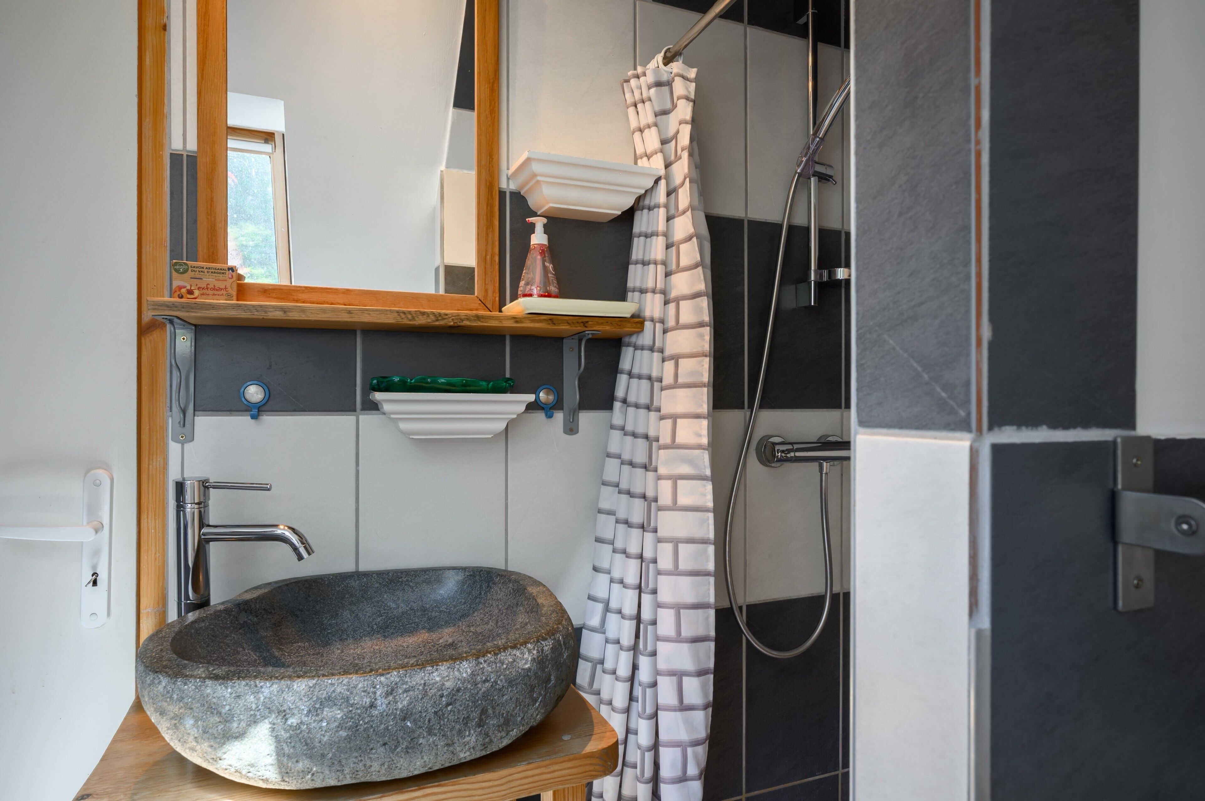 Ein Badezimmer mit einem Becken aus Stein, einem Holzregal, einem Spiegel, einer Duschvorhang und einem Handtuchhalter.