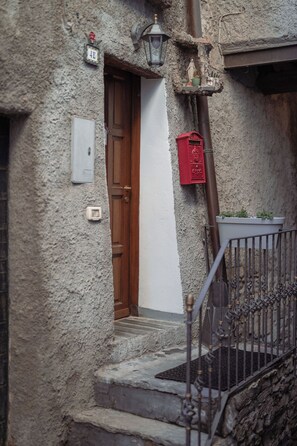 Uma escadaria de pedra estreita leva a uma porta de madeira, com uma caixa de correio vermelha e um candeeiro fixado na parede.