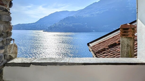 Ein Balkon mit Blick auf einen See und Berge, zwei Metallstühle und eine rustikale Steinmauer.