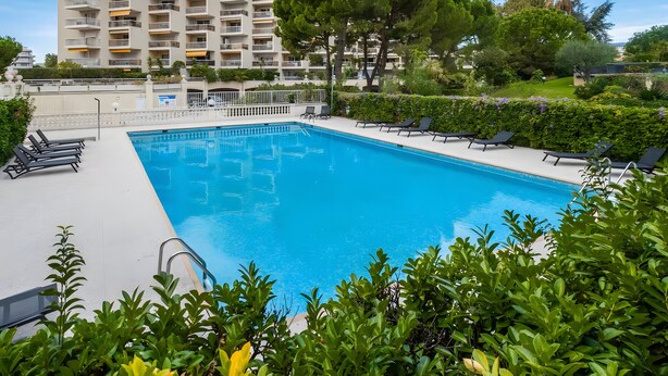 A swimming pool with lounge chairs, surrounded by greenery and a multi-story building in the background. - Le Lido Apartment – Beach, Pool, Parking, Sea View, Wi-Fi, Air Conditioning (Cagnes-sur-Mer)