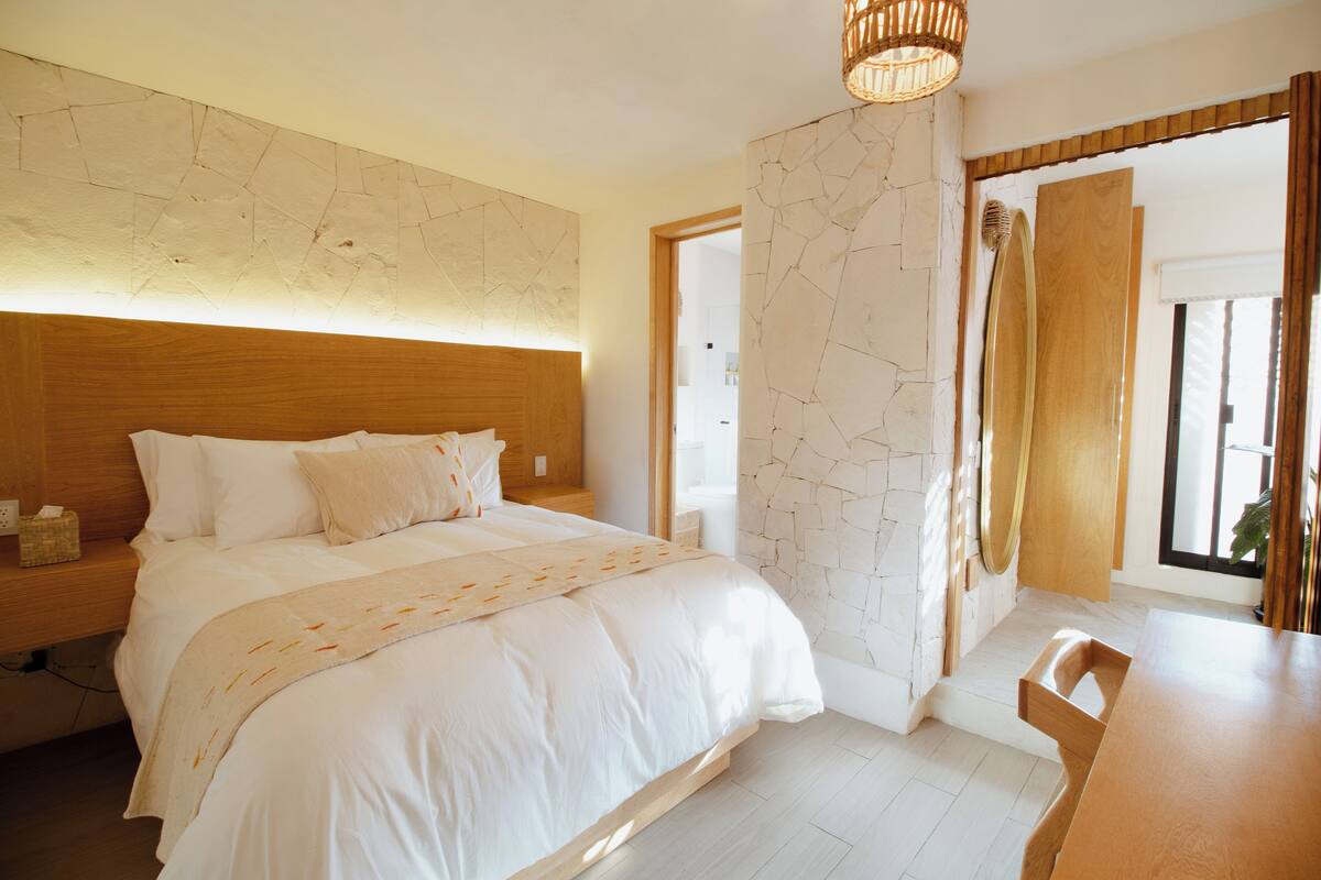 Ein modernes Schlafzimmer mit einem hölzernen Kopfteil, einem großen Bett mit weißer Bettwäsche, einem Nachttisch mit Taschentuchbox und einem Holzstuhl an einem Frühstückstisch.