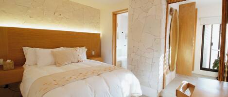 A modern bedroom with a wooden headboard, a large bed with white bedding, a bedside table with a tissue box, and a wooden chair at a breakfast table.