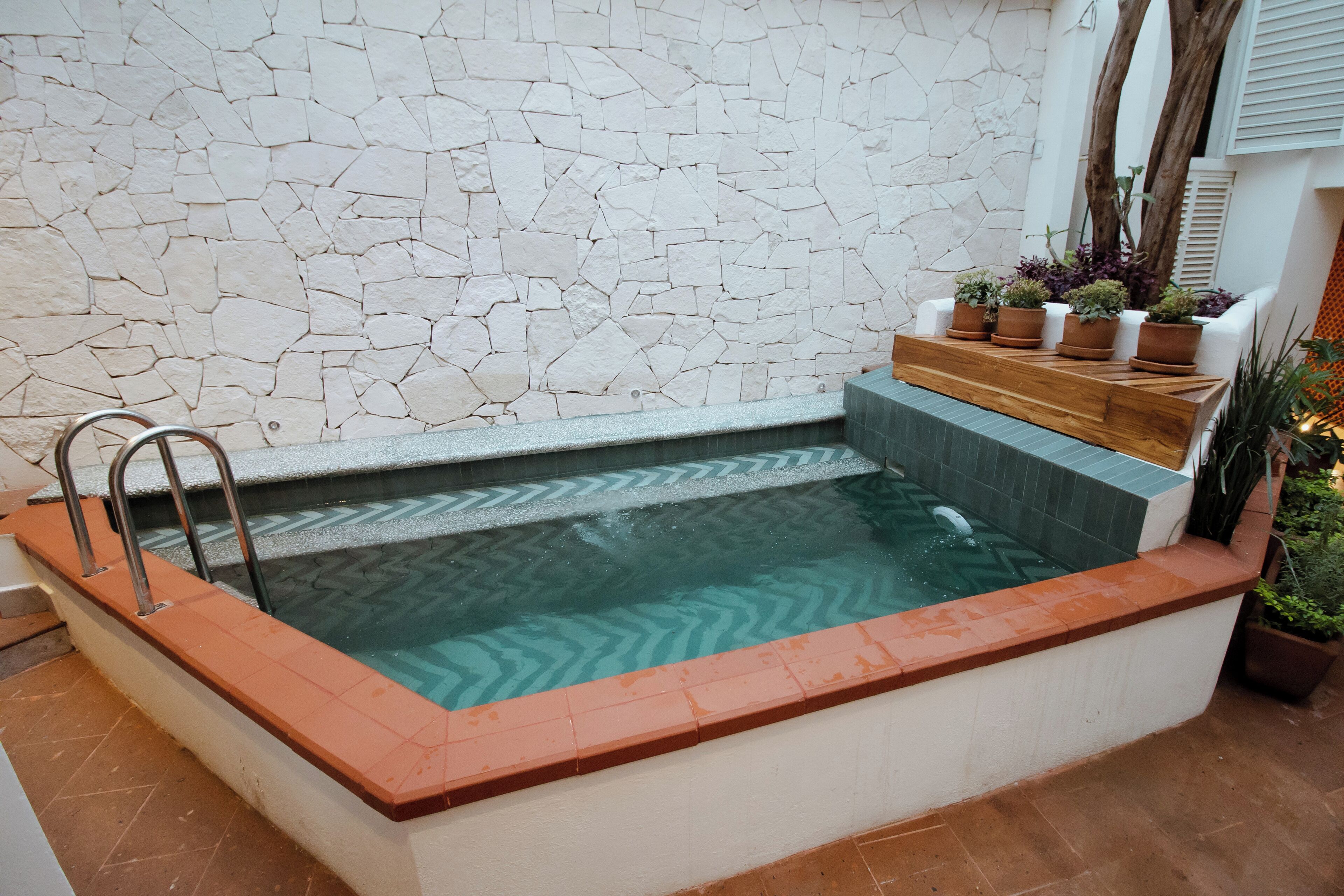 Una vasca idromassaggio rettangolare con uno sfondo a parete in pietra, un piano in legno con piante in vaso e una scala in metallo.