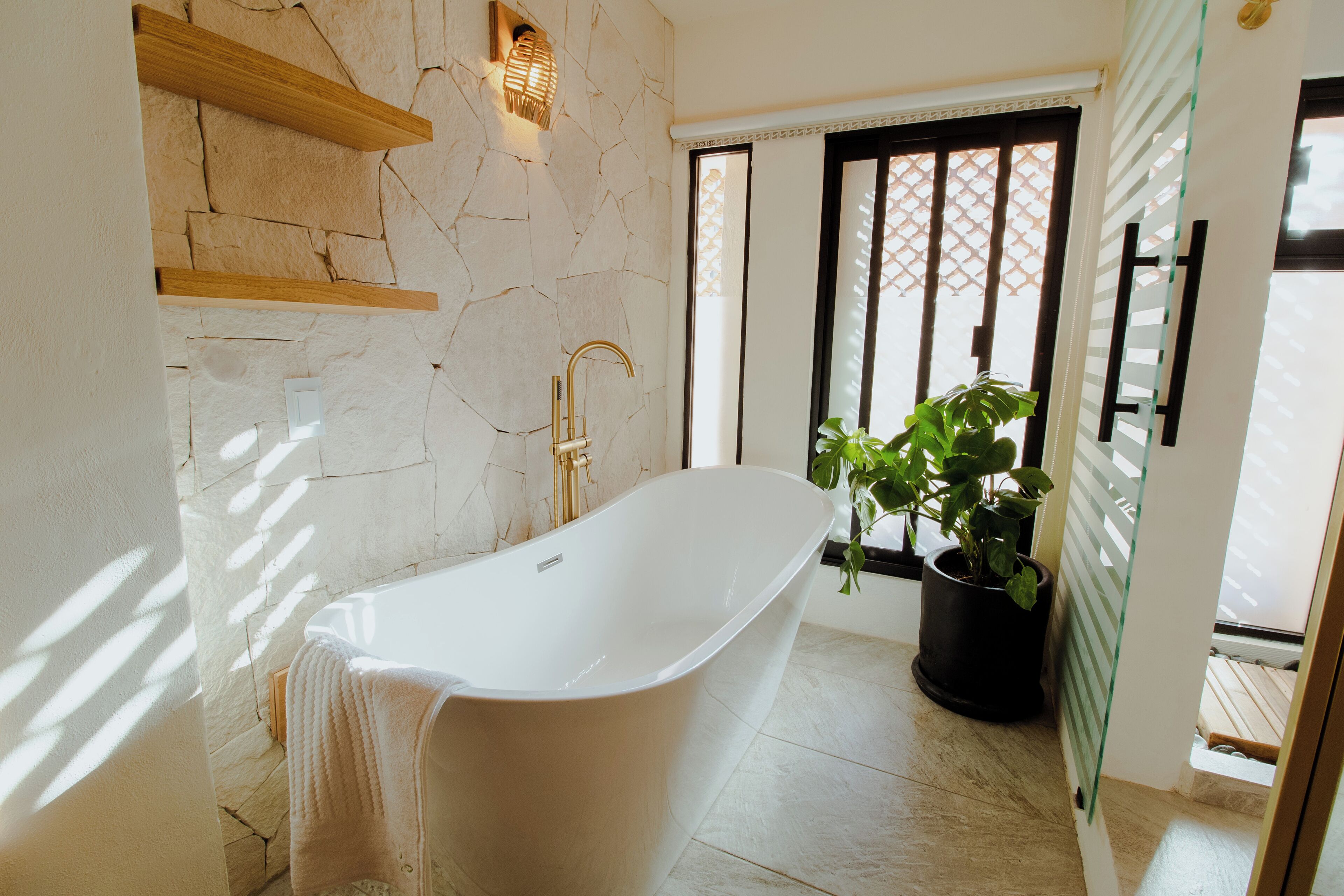 A modern bathroom with a freestanding bathtub, stone wall, wooden shelves, a potted plant, and a window with blinds.