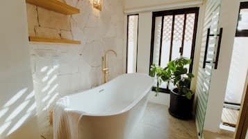 A modern bathroom with a freestanding bathtub, stone wall, wooden shelves, a potted plant, and a window with blinds.