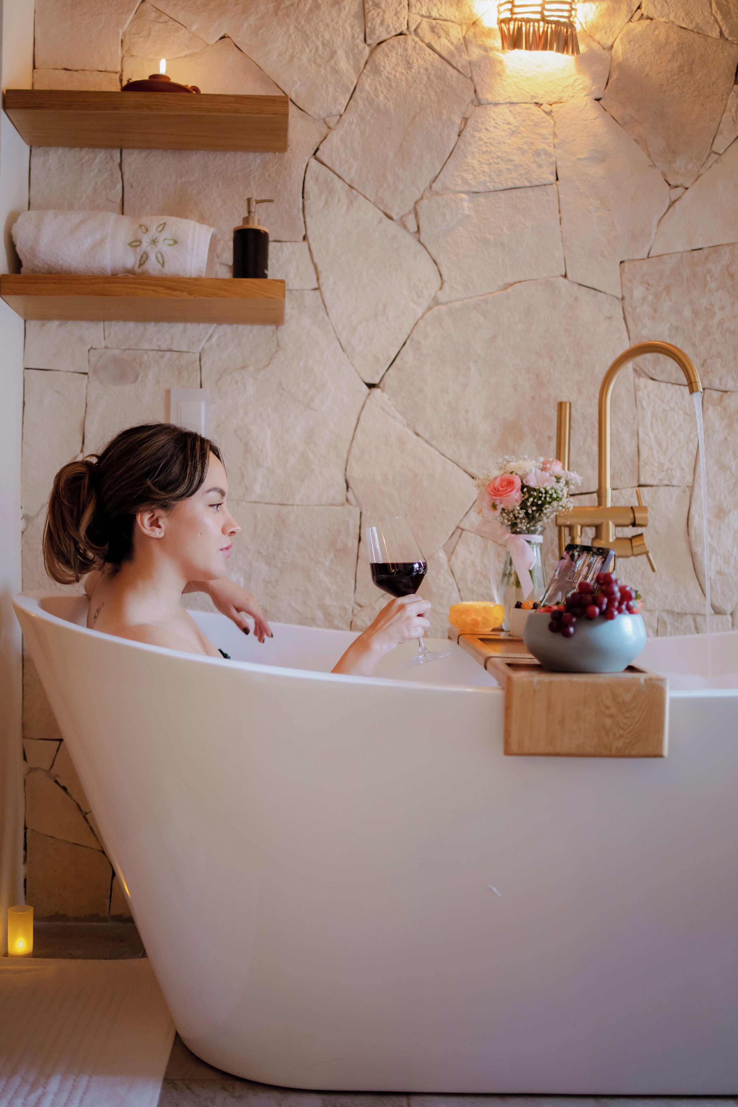 A woman in a bathtub with a bottle of wine, a bowl of fruit, and a vase of flowers.