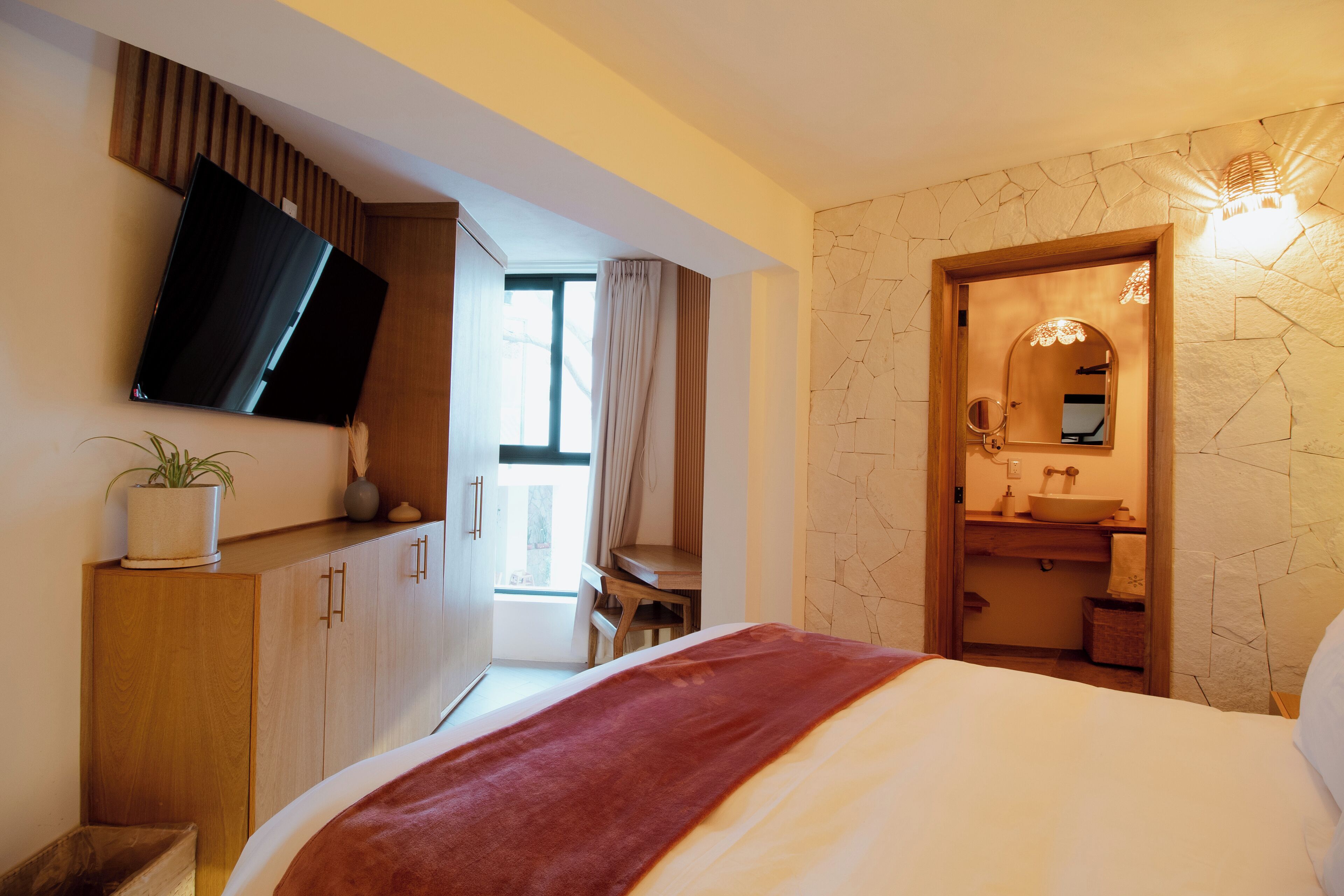 A modern bedroom with a bed, a TV mounted on the wall, a wooden desk, and a bathroom with a stone wall and a mirror.
