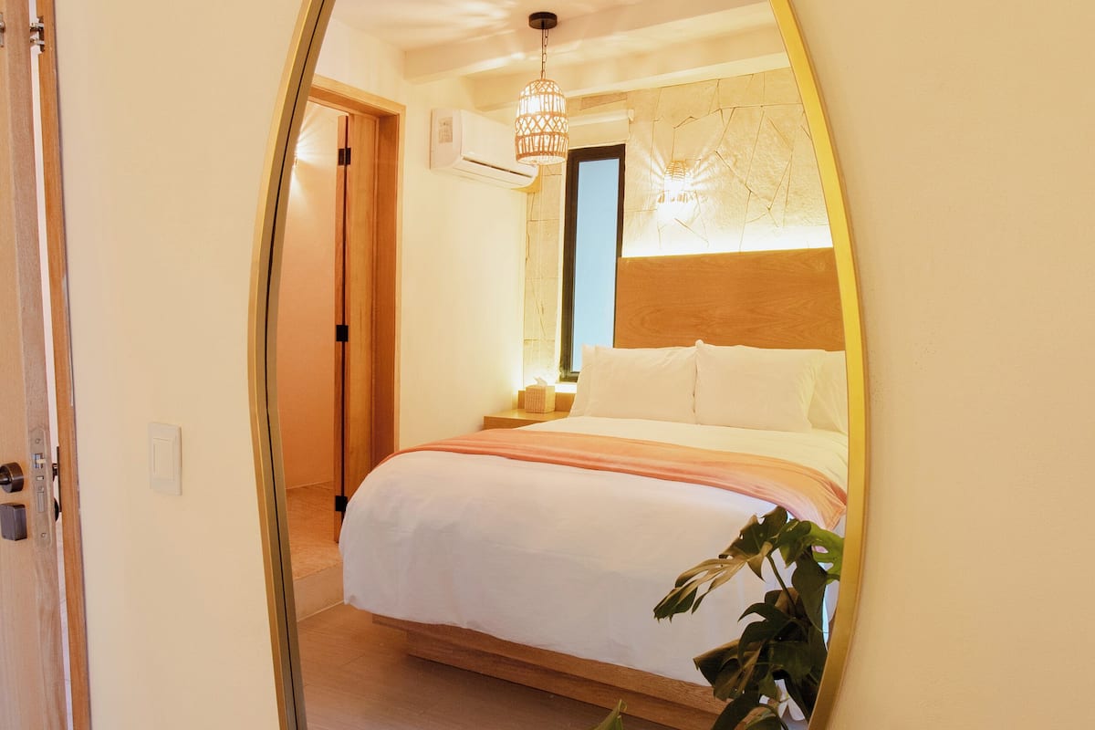 Ein Schlafzimmer mit einem großen Spiegel, der den Raum widerspiegelt, einem Bett, einem Nachttisch und einer Zimmerpflanze.