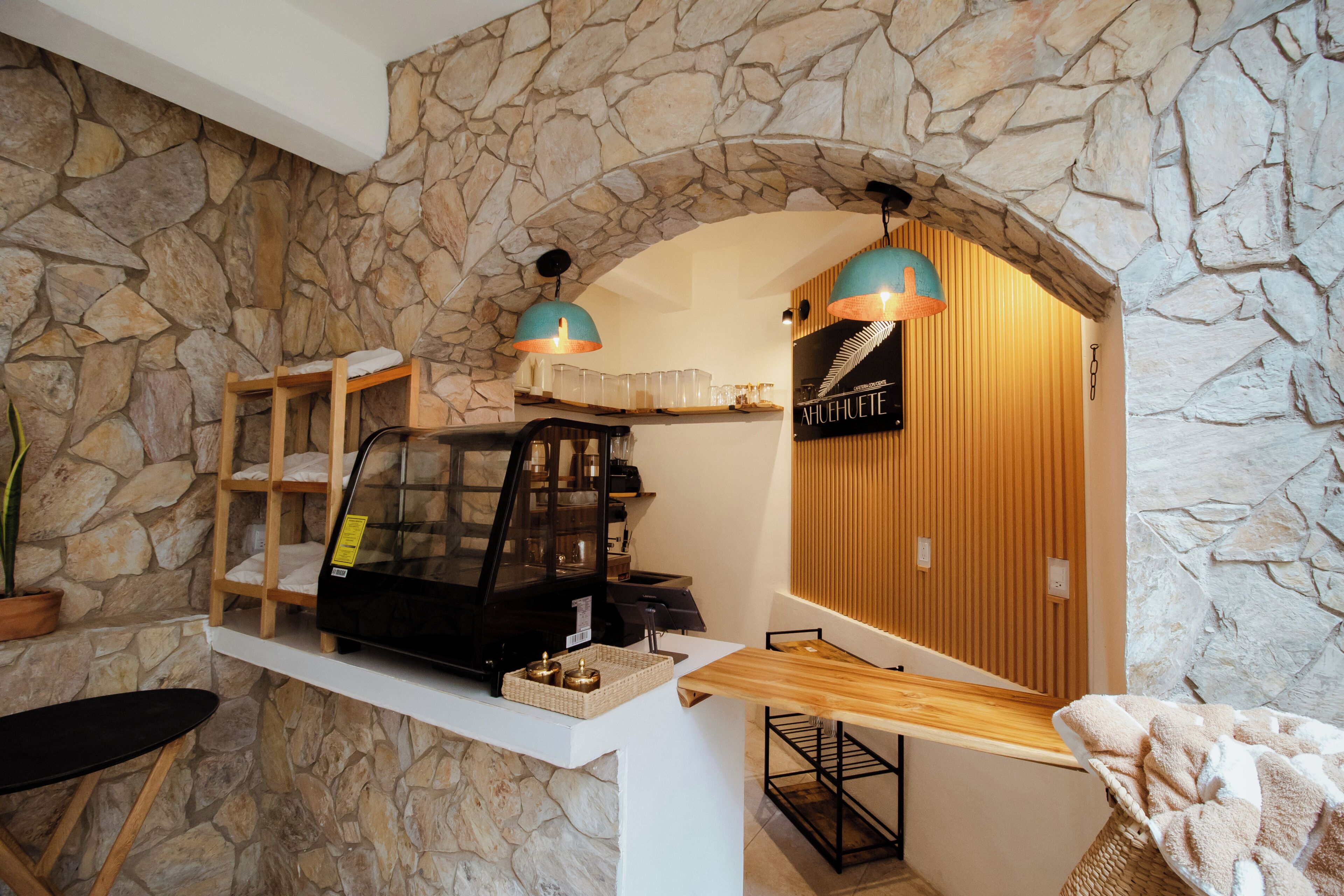 Una cafeteria amb paret de pedra, prestatges de fusta, una màquina de cafè i dues làmpades penjants.