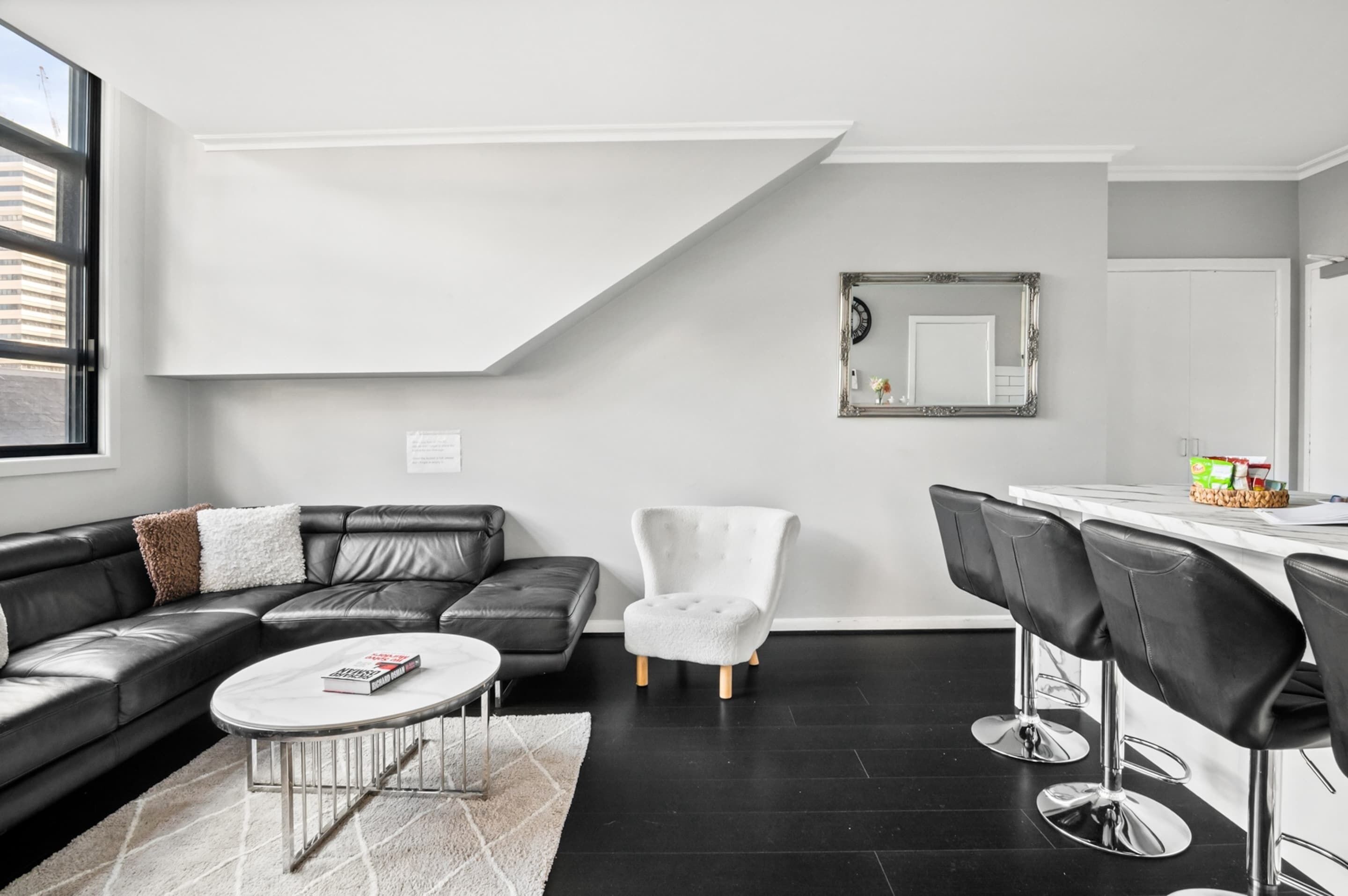 Una sala moderna con un sofá de cuero negro, un sillón blanco, una mesa de centro redonda y un comedor con taburetes de bar.