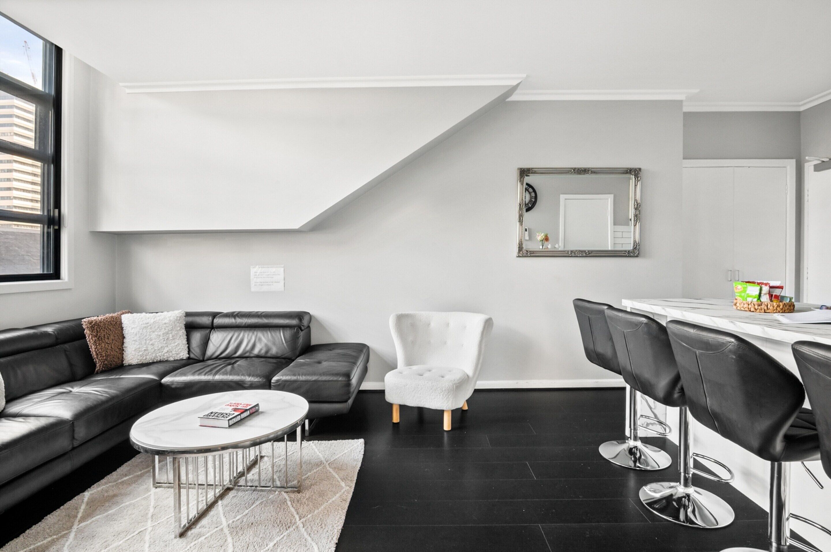 A modern living room with a black leather sofa, a white armchair, a round coffee table, and a dining area with bar stools.