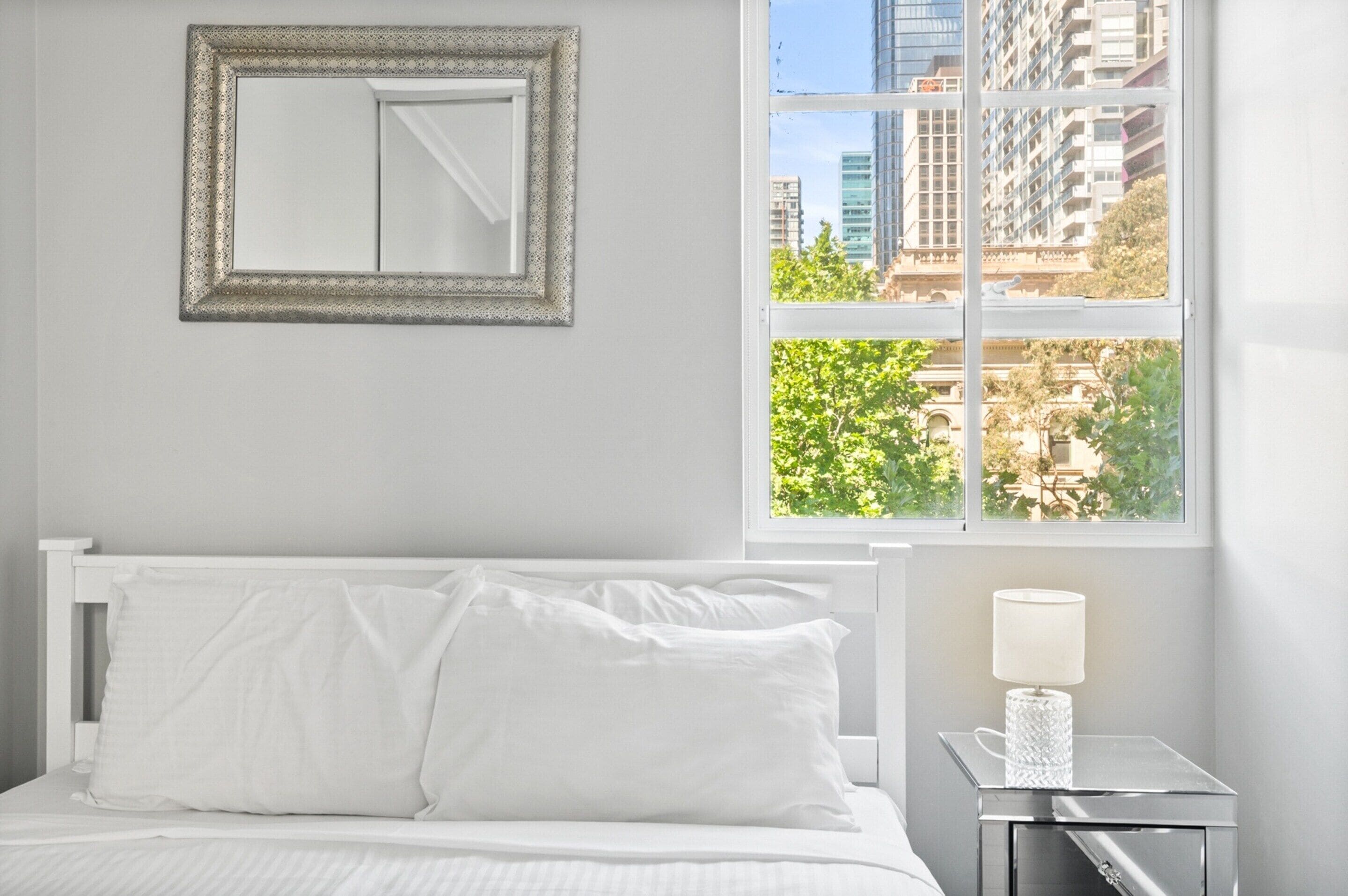 A hotel room with a bed, a nightstand with a lamp, a framed mirror, and a window with a view of buildings and trees.