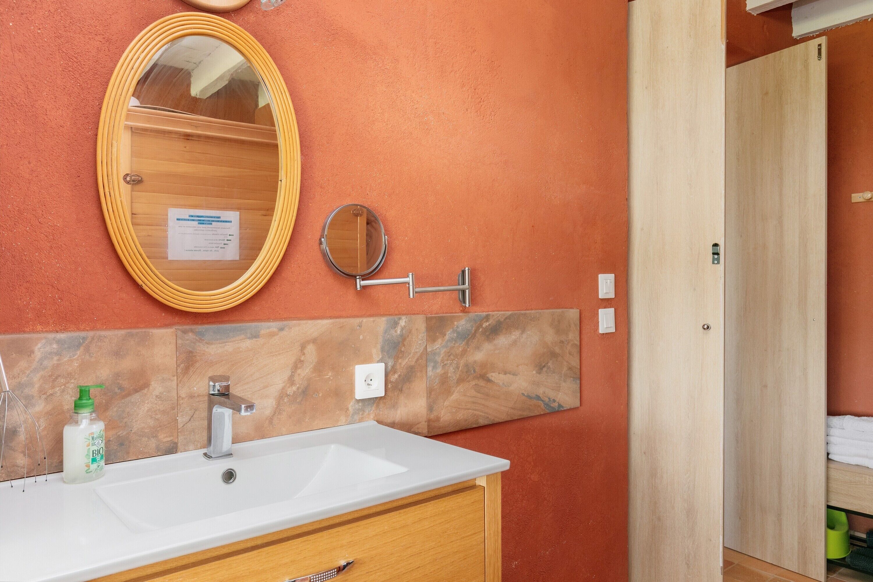 Une salle de bain avec un lavabo blanc, un meuble-lavabo en bois, un miroir rond et une porte en bois.