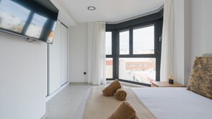 Ein Hotelzimmer mit Bett, Wandfernseher, Fenster mit Vorhängen und Blick auf Gebäude.