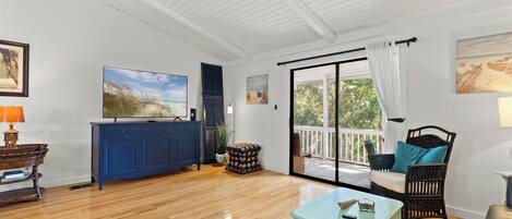 A spacious living room with a wooden floor, a blue cabinet, a television, and a balcony with a view of greenery.