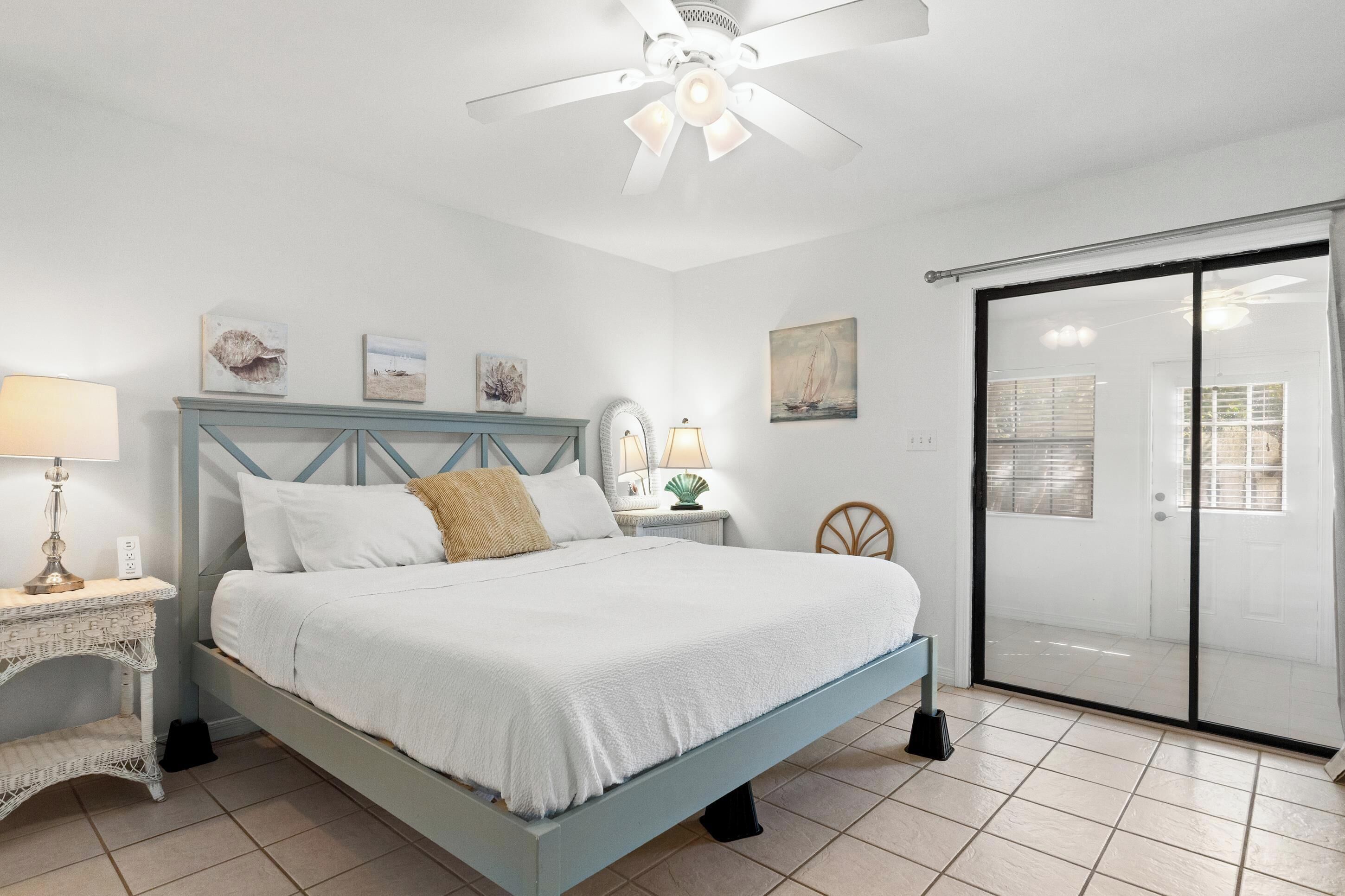 Quarto com cama, mesa de cabeceira, luminária, ventilador de teto e porta de vidro deslizante.