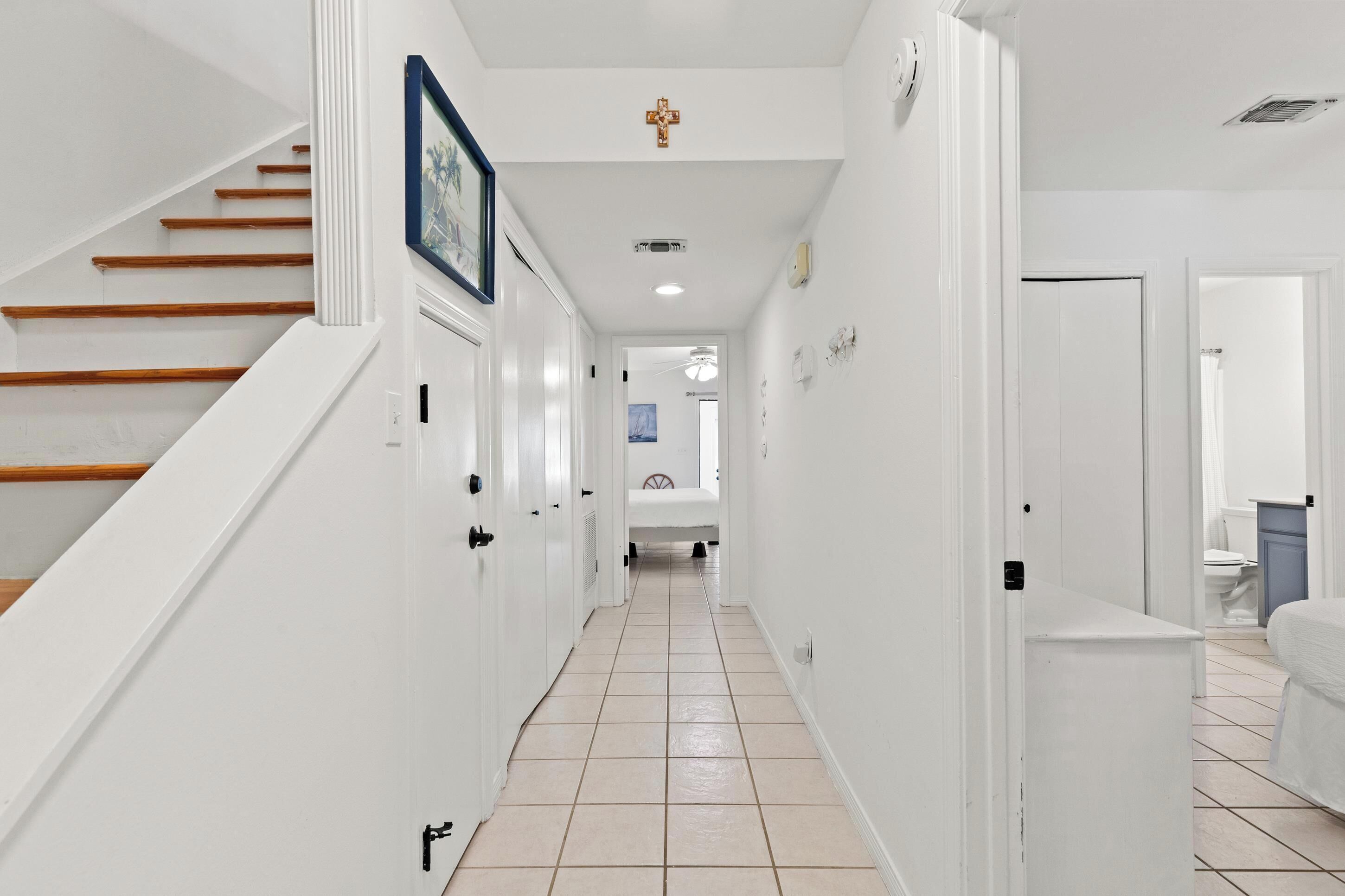 A narrow hallway with white walls, a wooden staircase on the left, and a bathroom visible at the end.