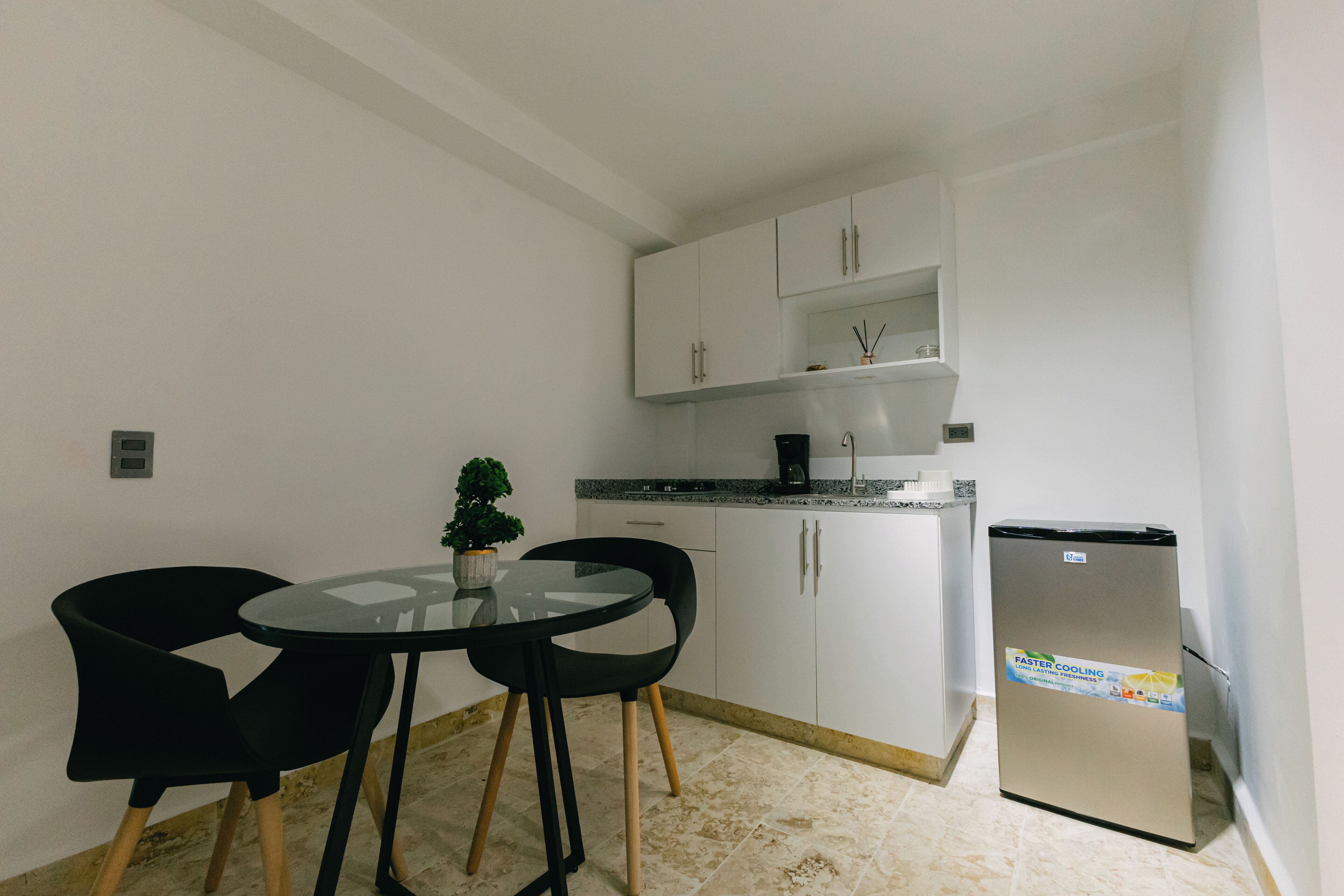 Une cuisine compacte avec des meubles blancs, un réfrigérateur, une petite table à manger avec deux chaises et une plante en pot.