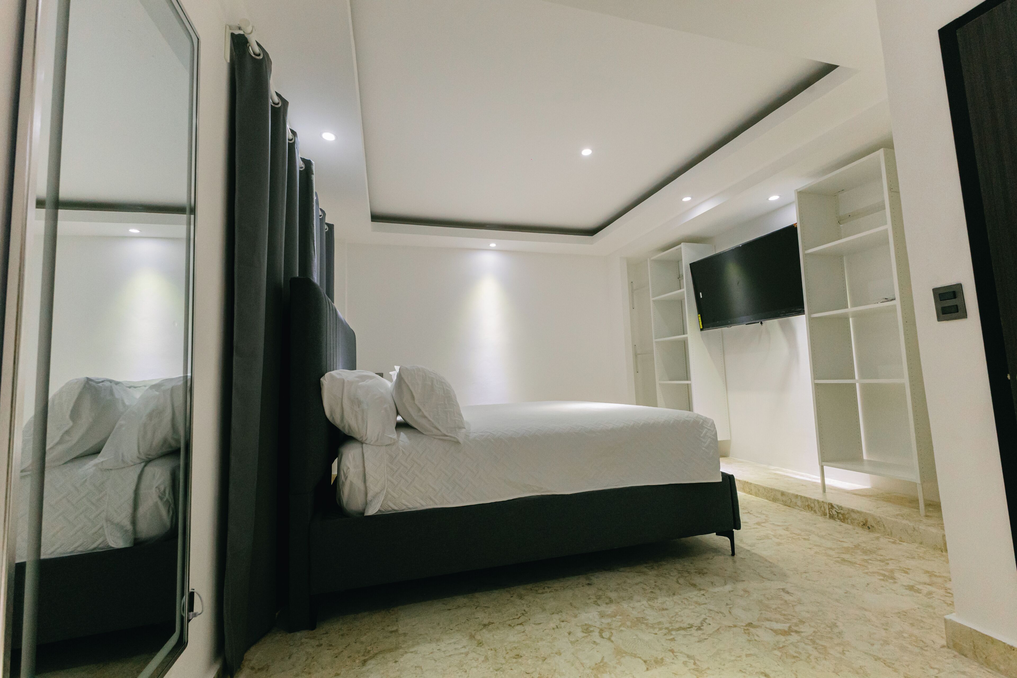 A modern bedroom with a bed, a large mirror, a TV, and built-in shelving.