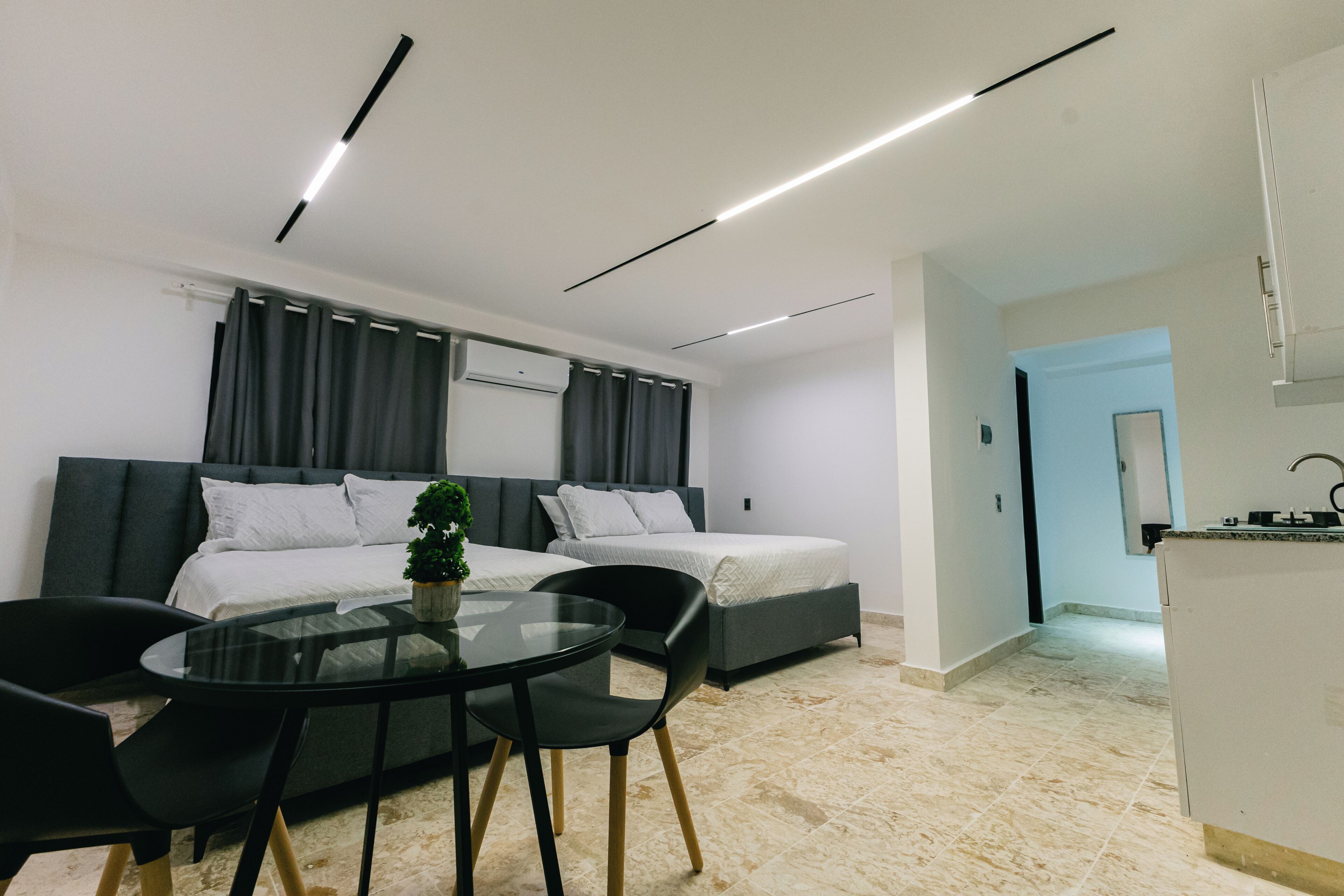 A modern hotel room with two beds, a dining area with a glass table, and a kitchenette.