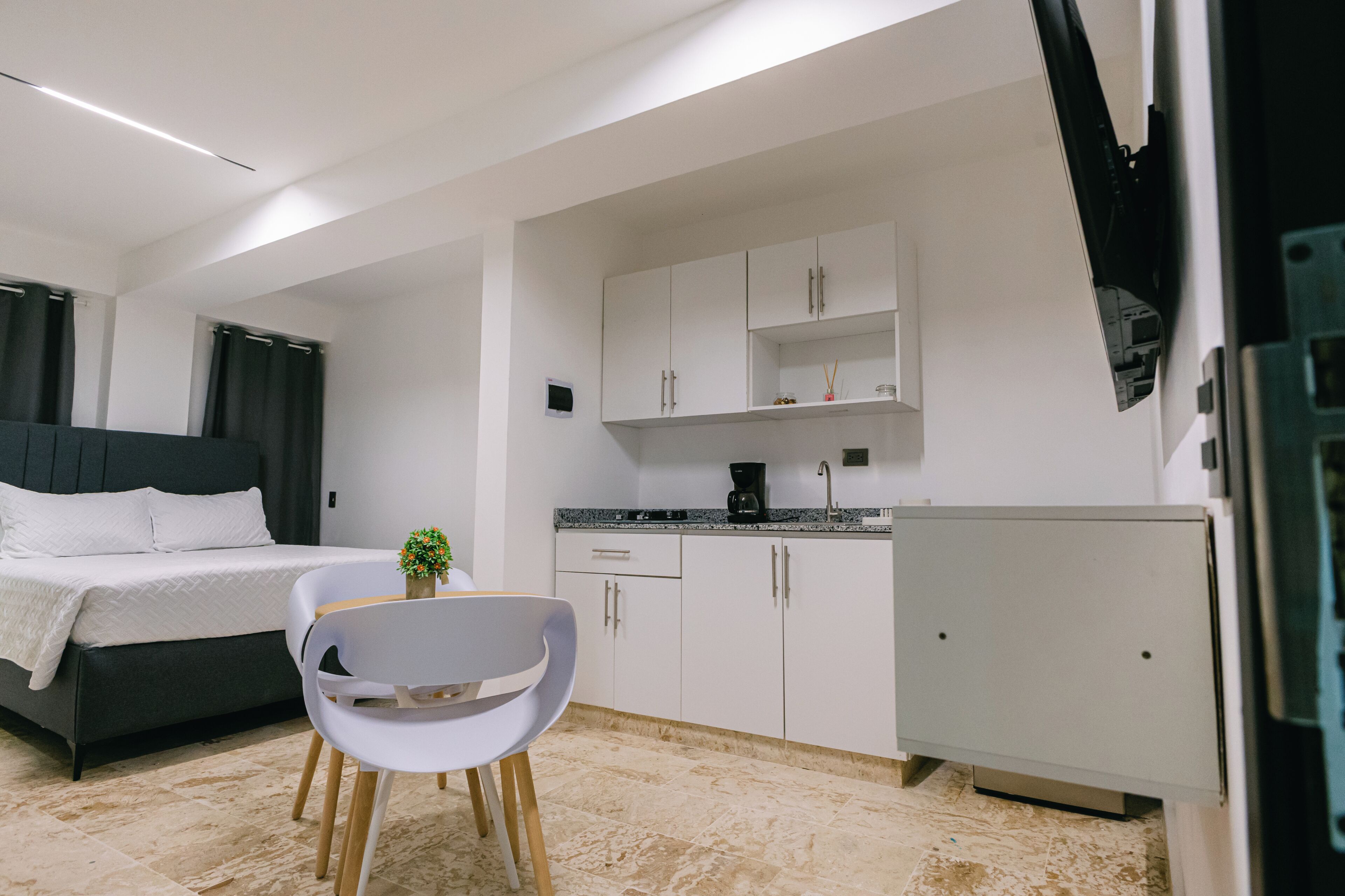 A modern bedroom with a bed, a small dining area with a chair, and a kitchenette with cabinets and a coffee maker.